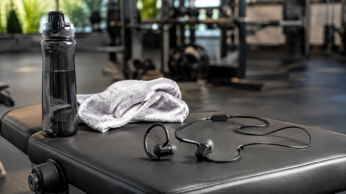 Unbranded workout headphones beside a towel and water bottle on a gym bench